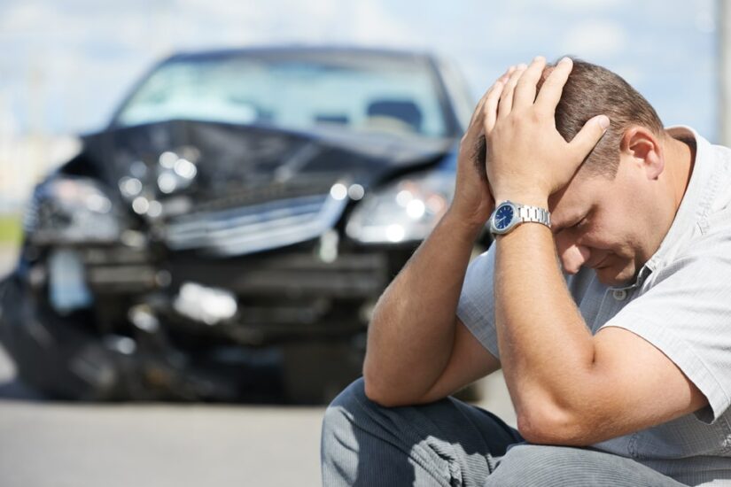 Man Holding His Head Next To A White Damaged Car